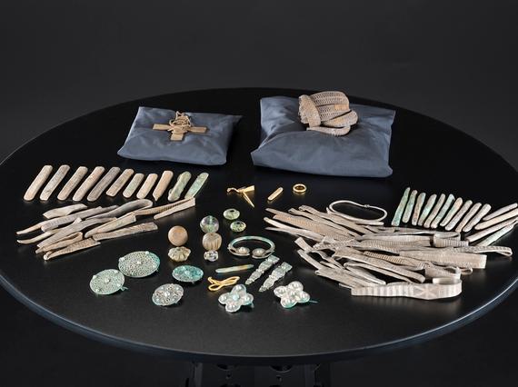 Several silver objects are laid out on a black table. Pillows prop up a cross and arm-ring bundle with the rest organised by object type.