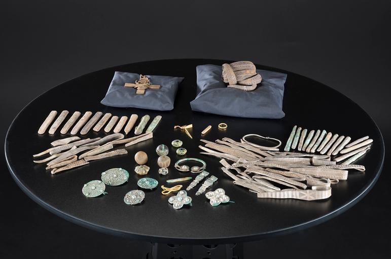 Several silver objects are laid out on a black table. Pillows prop up a cross and arm-ring bundle with the rest organised by object type.
