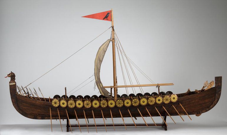 A model ship of a long wooden viking boat with oars and shields with a red flag on top