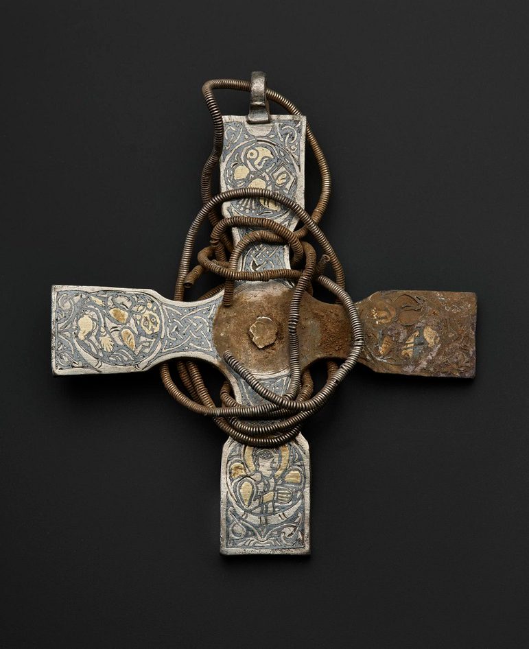 A silver and gold viking-age cross with a chain coiled around it. The left arm of the cross is covered dirt.