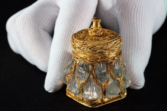 Light green crystal jar with gold base, lid and mesh-like covering against a black background with hand for scale. Jar is no taller than a finger.