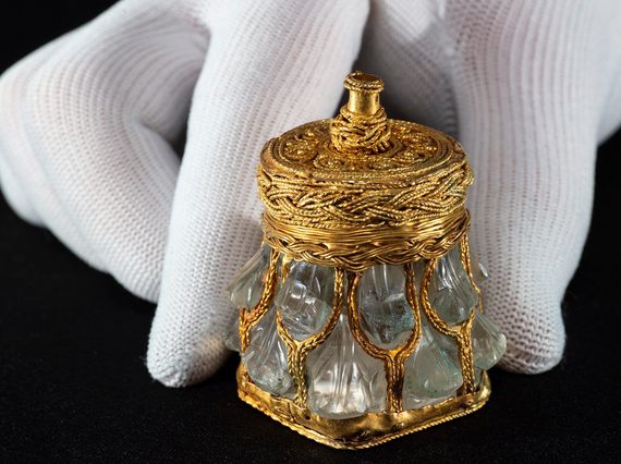 Light green crystal jar with gold base, lid and mesh-like covering against a black background with hand for scale. Jar is no taller than a finger.