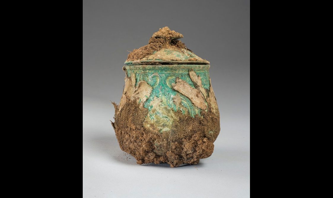 A faded green lidded pot with tufts of brown fabric attached to its surface