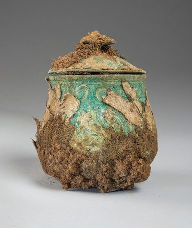 A faded green lidded pot with tufts of brown fabric attached to its surface