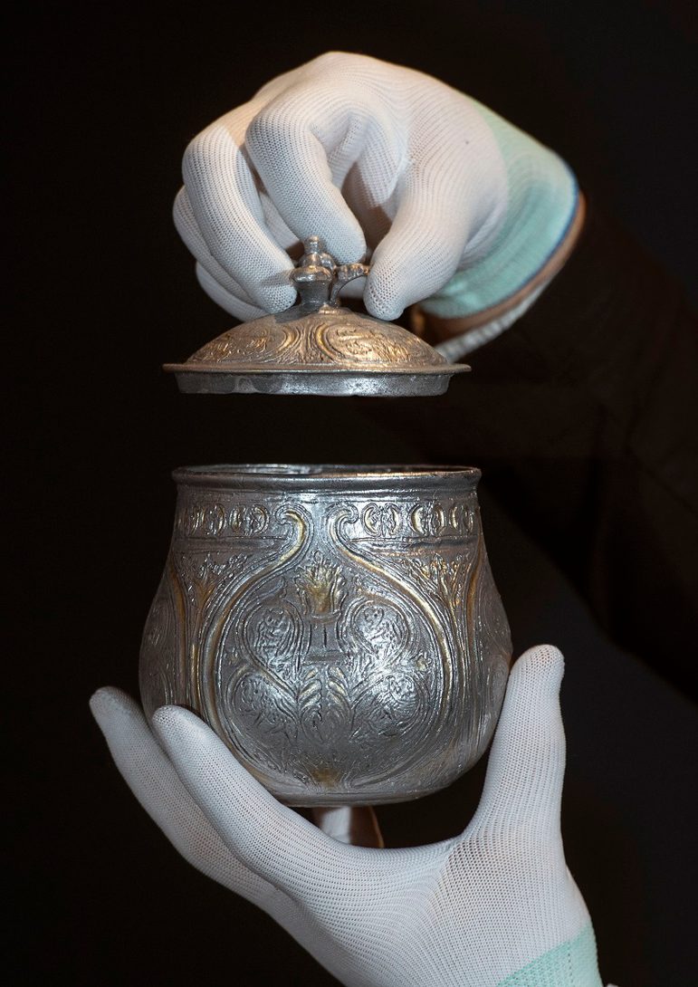 A silver round vessel with a gilded lid engraved with various round and organic shapes, held in the air by a set of hands set against black background.