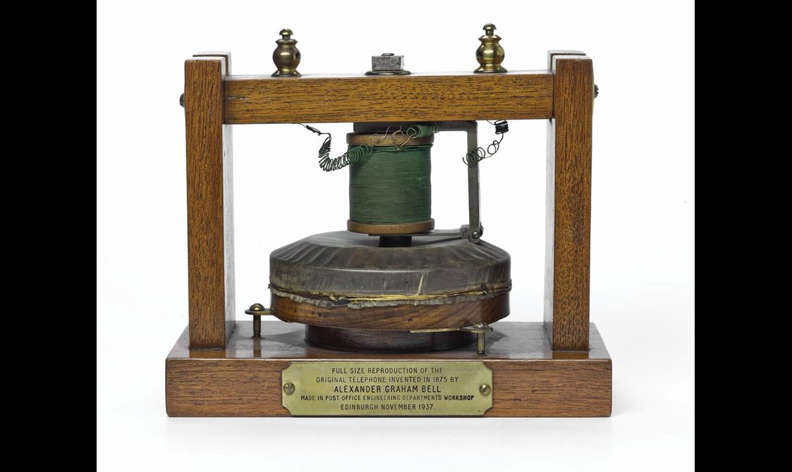 An early telephone which has a wooden frame and a round object sitting on the base with a spool with wire on top.