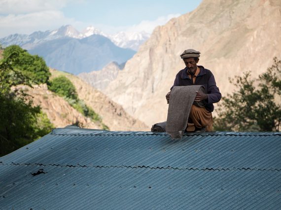 Man sitting on a corrugated metal roof rolling a length of brown fabric. There are large, snowcapped mountains in the background.