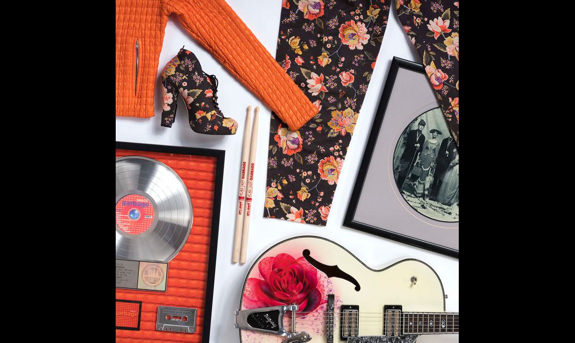 Objects belonging to the artist Garbage, including an orange jacket, floral shoes and trousers, a pair of drumsticks, a black and white photograph of the band, and a framed vinyl record.