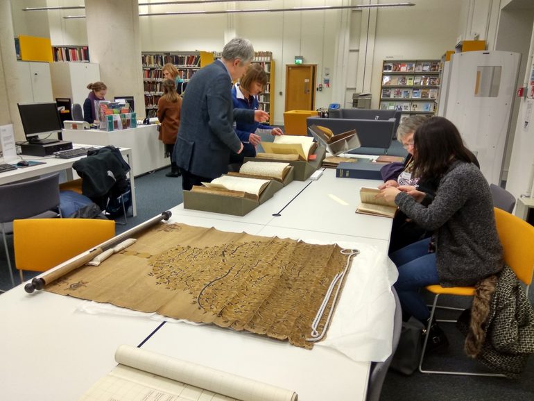People sit and stand around a large white table holding old books and a large scroll with a tree-like shape on it.