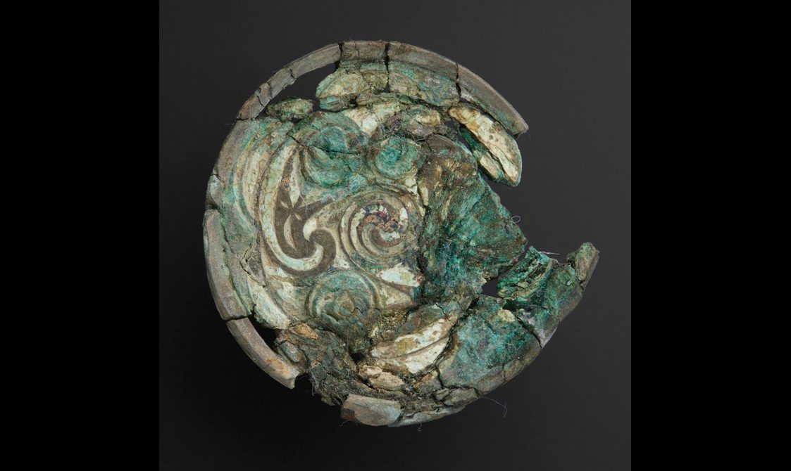 A disc that has been damaged in grey, green colours, with Celtic style patterns on its surface