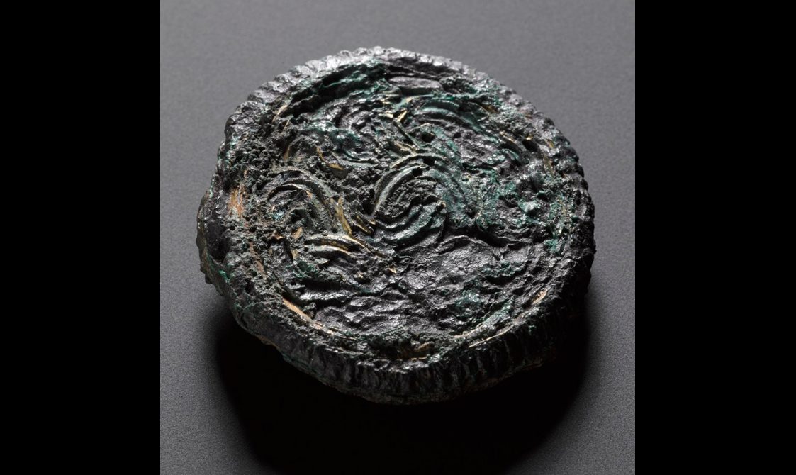 A mottled round object in brown and green colours with celtic like details on its surfaced