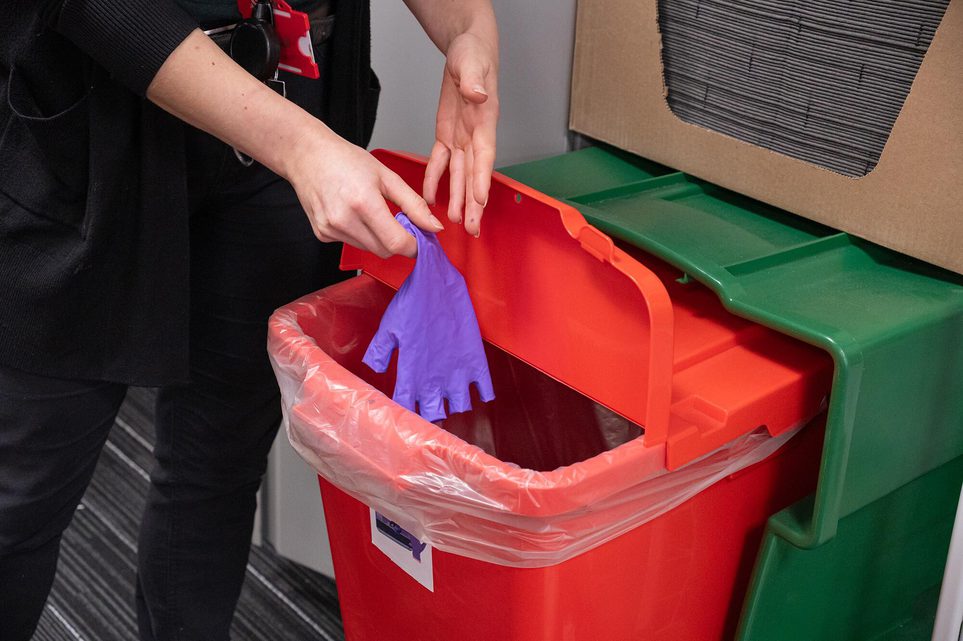 A curator putting a pair of purple nitrile gloves into a red recycling box.