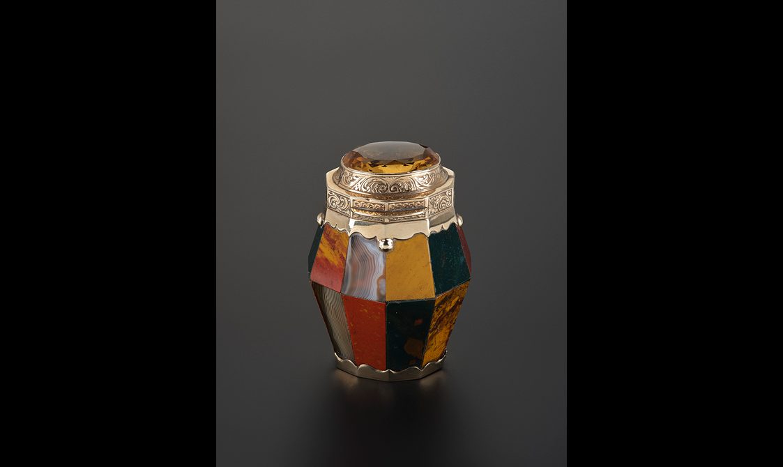 A small faceted jar made with agate and gold. A yellow cairngorm sits atop the lid.