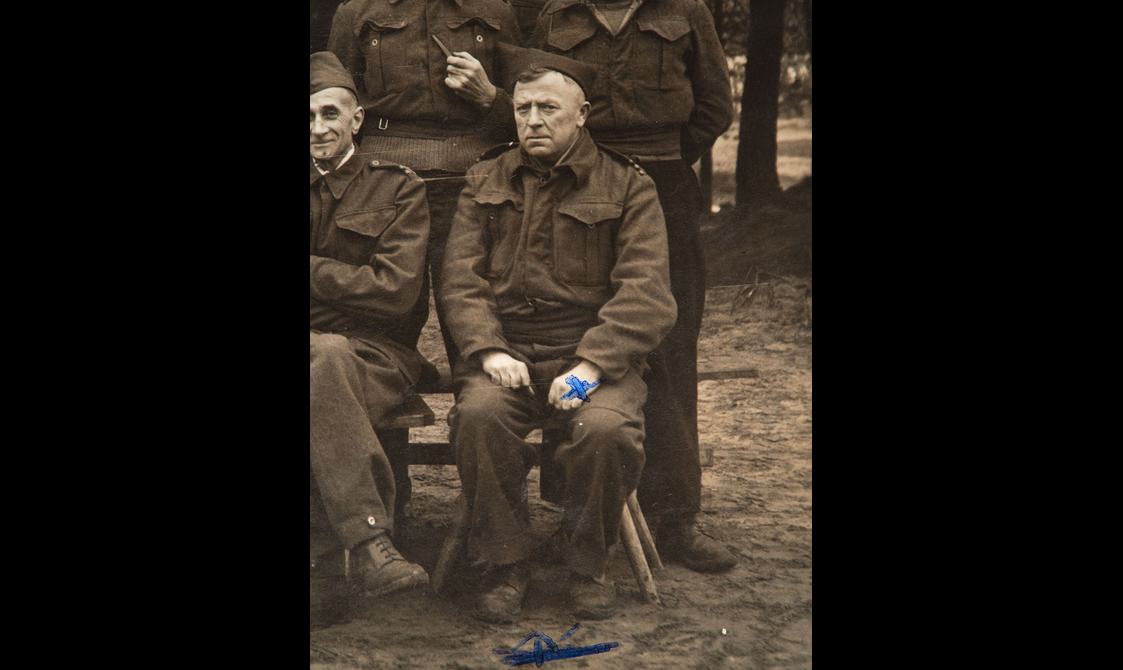 An army lieutenant in uniform sitting down for a group photo. There is a blue cross marked on his hand.