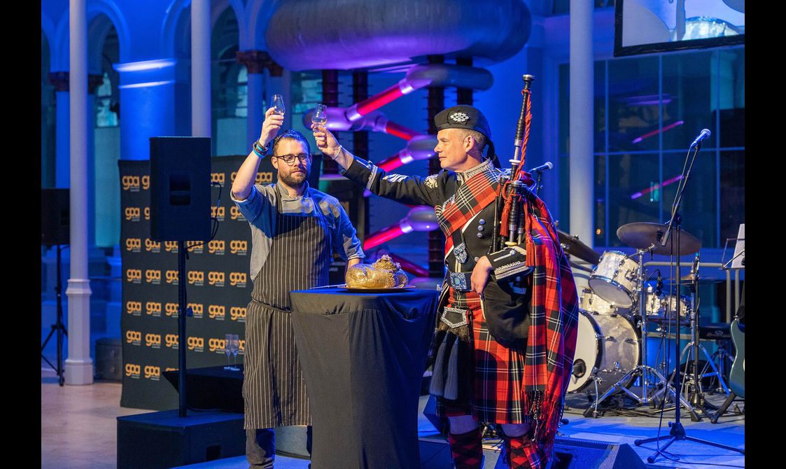 A piper and a chef raising glasses of whisky to toast a haggis at a Burns Night event.
