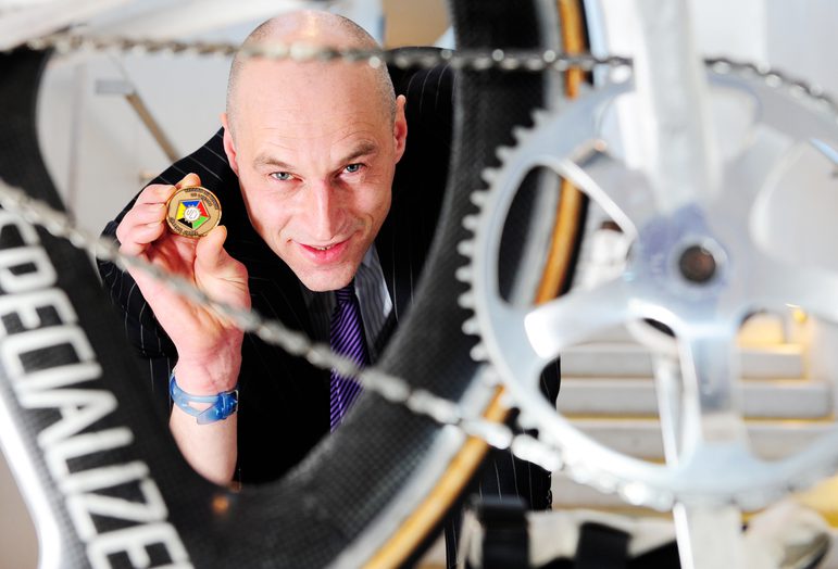 A bald man wearing a suit holding a gold and multicoloured medal behind the spokes of a bike wheel