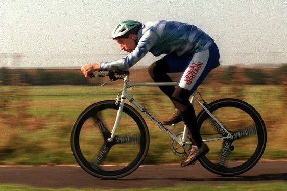 A man cycling a bicycle with his back low, looking forward. There are green fields in the background