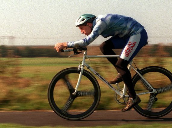 A man cycling a bicycle with his back low, looking forward. There are green fields in the background