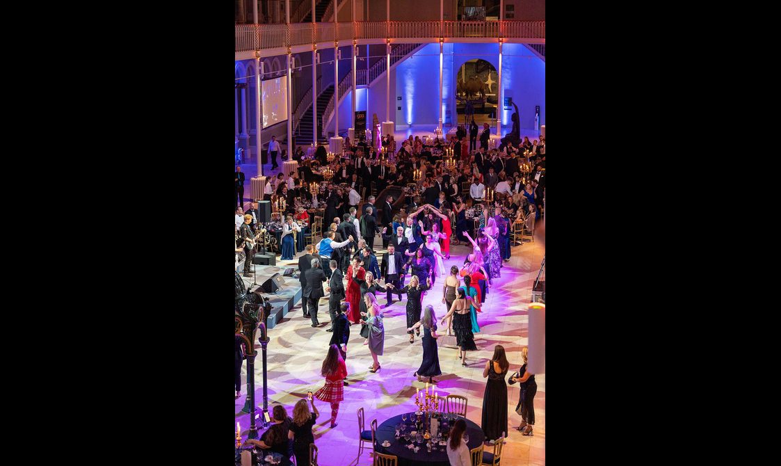 People dancing at a ceilidh in a large museum gallery. There is a band playing and dinner tables lining the dancefloor.