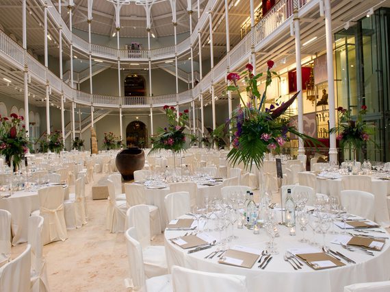 A large, bright, multistory space with balconies running around the sides. The floor is filled with large round tables covered in white tablecloths and set for a meal. The tables are surrounded by white chairs.