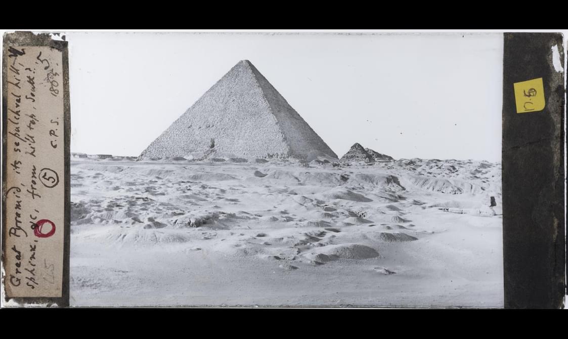 A black and white image of a pyramid in a sandy landscape. There is a yellowing label to the left of the image with handwritten notes scrawled on it.