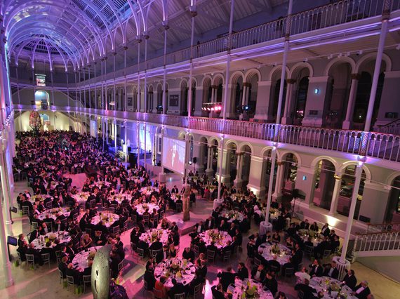 A large, multistorey gallery space with balconies running around the outside. Low, blue and pink lighting illuminates a floor of round tables full of people.