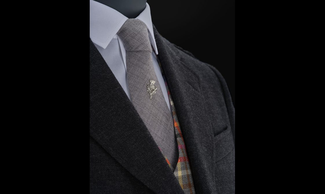 A close up of a mannequin wearing a white shirt, dark jacket, and colourful tartan waistcoat with a grey woollen tie with a thistle pin.