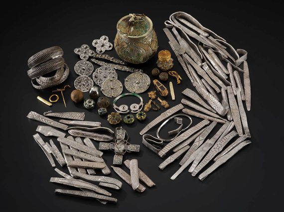 A large selection of old objects including armbands and brooches made of silver, decorative stones, and a lidded jar all sit arranged against a black background.
