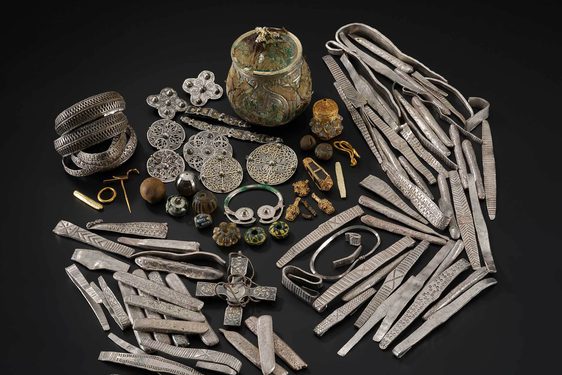 A large selection of old objects including armbands and brooches made of silver, decorative stones, and a lidded jar all sit arranged against a black background.