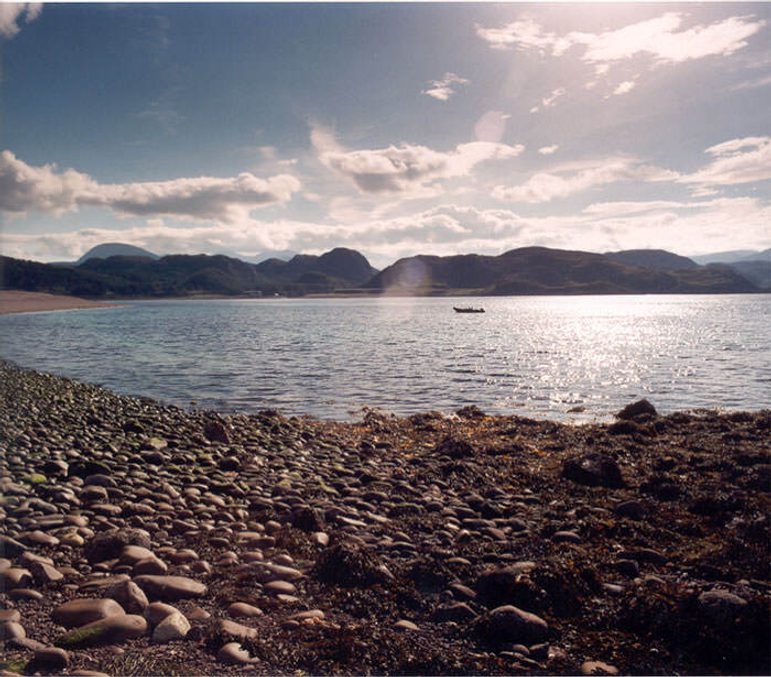 A small bay on a sunny day, with a rocky beach in the foreground and mountains in the distance. There is a small boat in the bay.