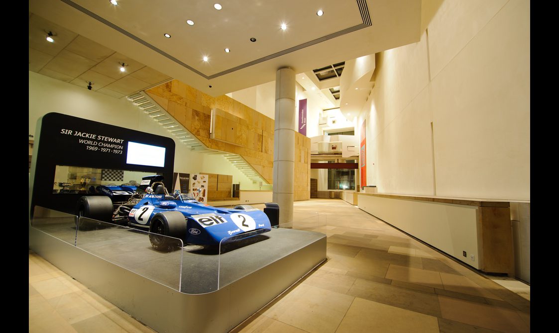 A large museum space with stone floors, high ceilings, and white walls. There is a formula one racing car on a platform.