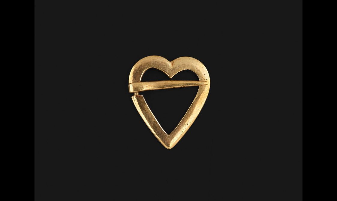 A gold heart brooch with a long pin going across the top of the heart