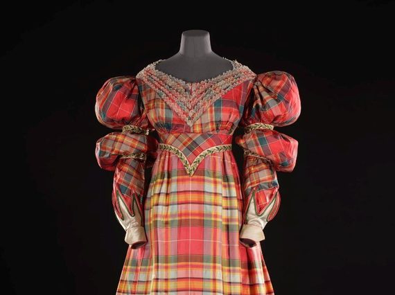A mannequin wearing an elaborate dress made of red silk tartan. The dress has long sleeves with puffy shoulders and is fitted at the waist.
