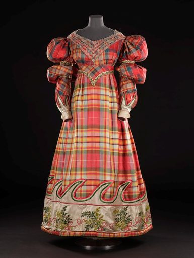 A mannequin wearing an elaborate dress made of red silk tartan. The dress has long sleeves with puffy shoulders and is fitted at the waist.