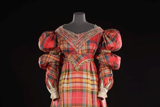 A mannequin wearing an elaborate dress made of red silk tartan. The dress has long sleeves with puffy shoulders and is fitted at the waist.
