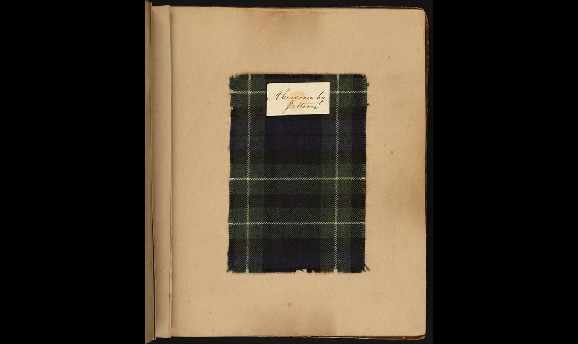 A swatch of a dark green checked tartan fabric. It is mounted on a yellowing page and has a label sitting at the top of the swatch.