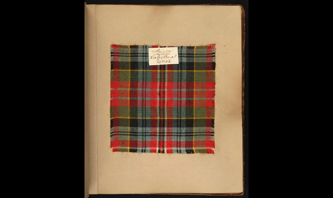 A swatch of a red, green, and yellow tartan fabric fixed to a piece of yellowing card with a label fixed to the top of the swatch.
