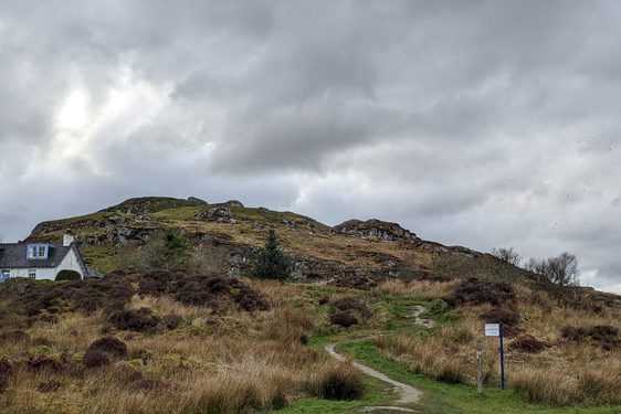 A rocky hill with with patches of grass and heather with a walking trail winding upwards. There is a small white house with a blue door and window frames to the left