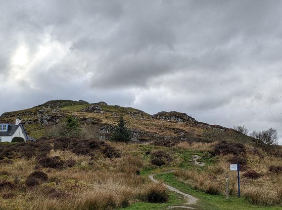 A rocky hill with with patches of grass and heather with a walking trail winding upwards. There is a small white house with a blue door and window frames to the left