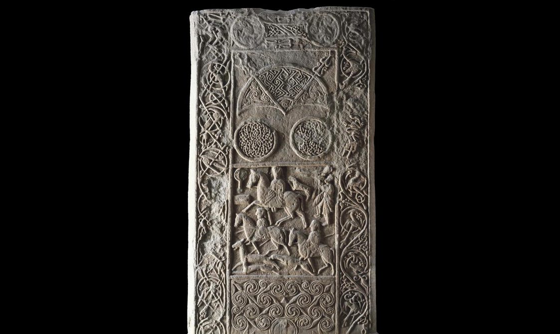 Intricately decorated stone with men riding horses in the middle and ornamental motifs on the sides.