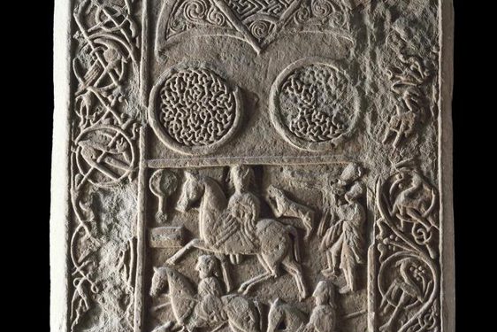 Intricately decorated stone with men riding horses in the middle and ornamental motifs on the sides.