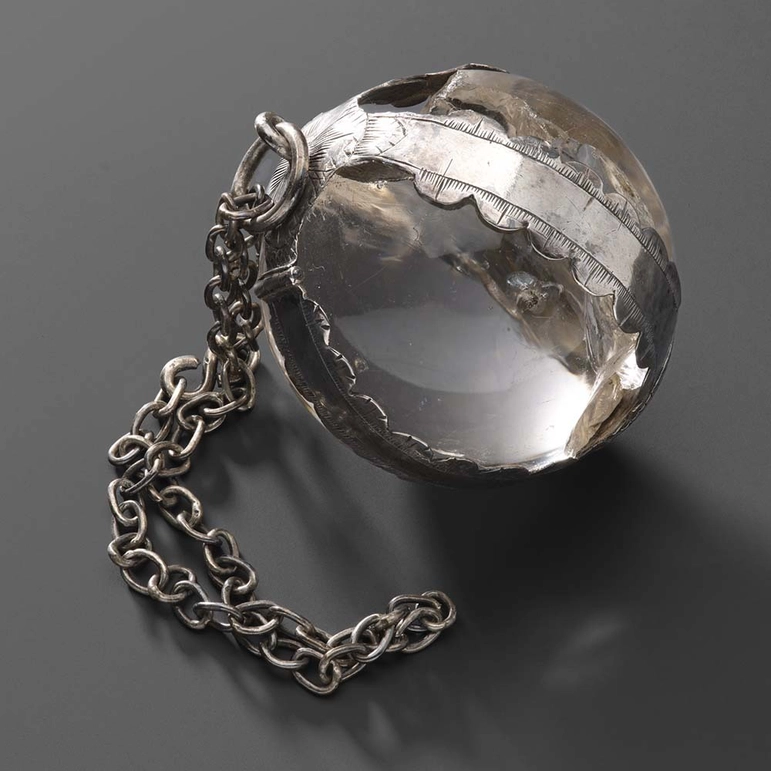 A transparent crystal ball held within a silver frame. The charmstone has a suspension loop, to which a chain is attached.