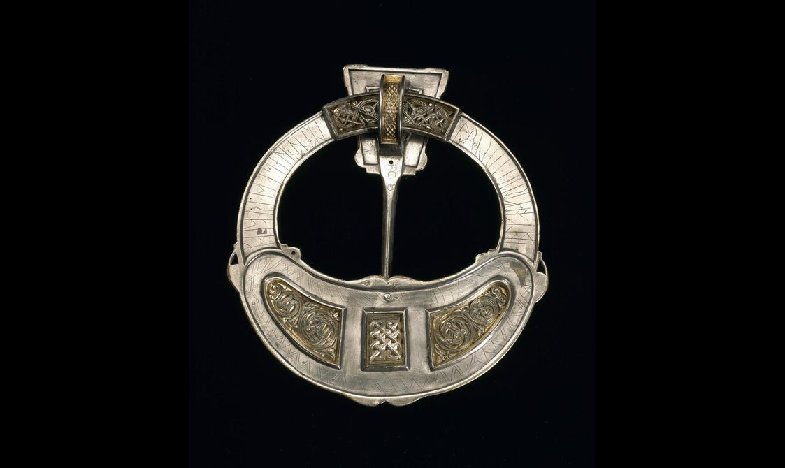 A circular brooch resembling a belt buckle, with light linear carvings around the rim and ornate panels of gold filigree.
