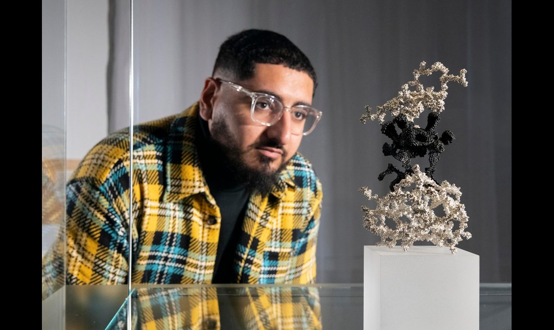 A man wearing glasses is peering into a glass museum case. Inside is a small sculpture showing an artistic representation of two white cells, and in between, a black one.