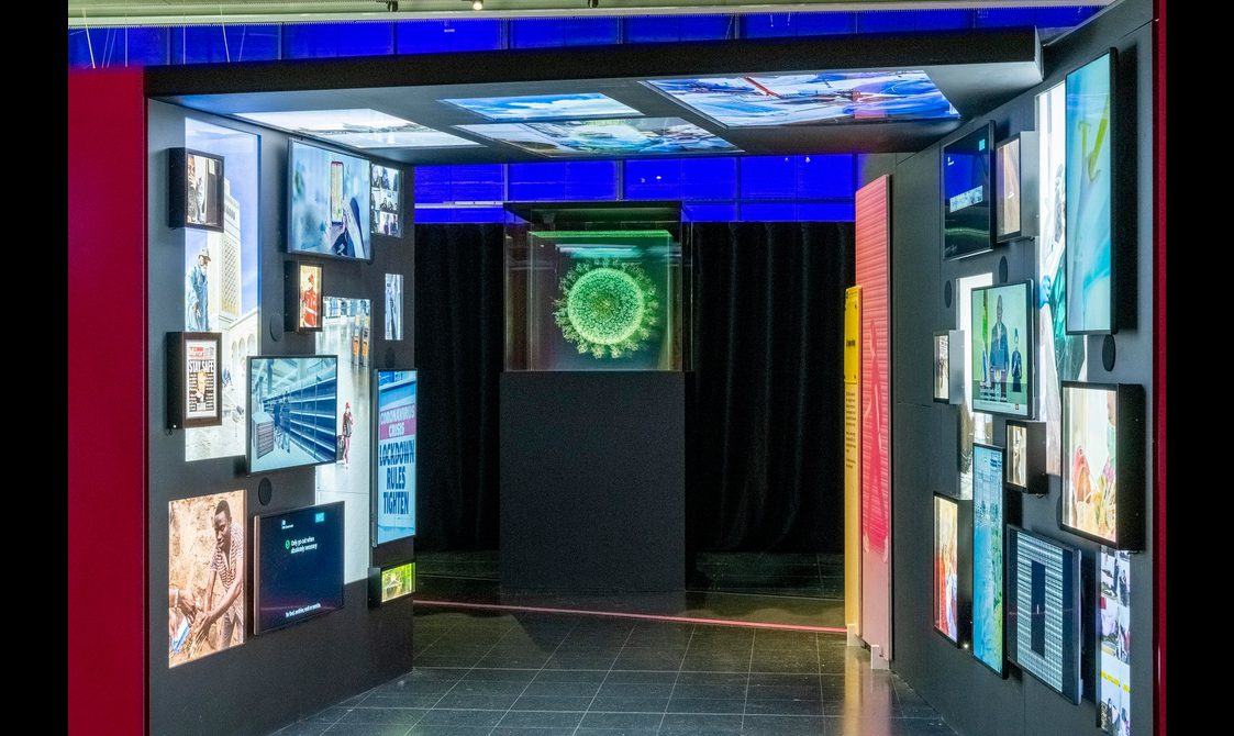 An exhibition display. At the centre of the image is an artwork on top of a black plinth. It looks like a light installation in the shape of a neon green virus cell.