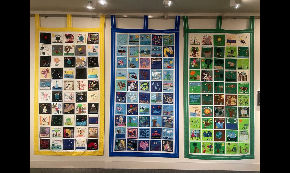 Three large vibrant quilts hanging against a white wall. Made up of colourful squares showing patterns, shapes and words of hope for the future.