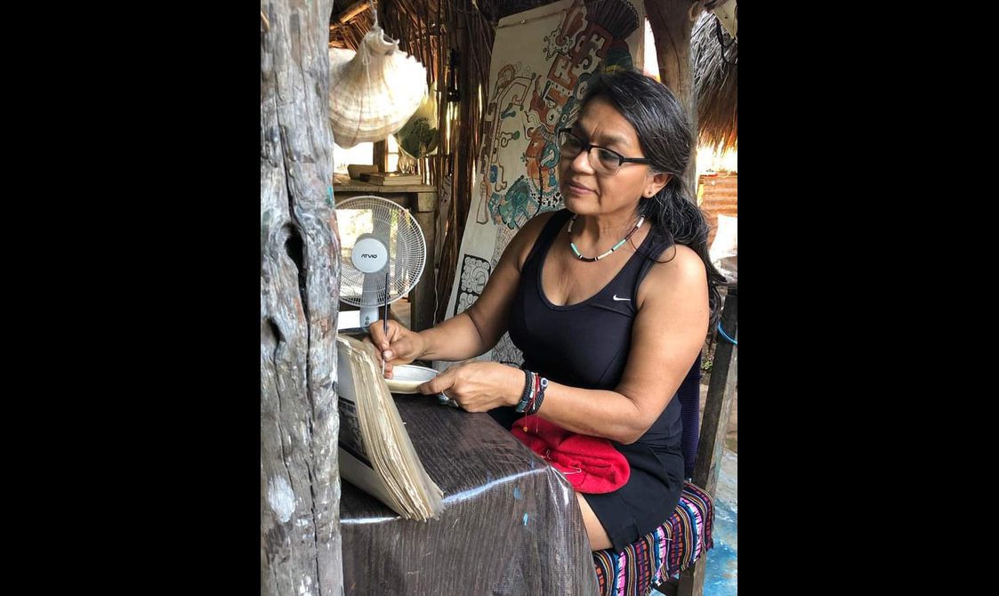 A woman is sat at a table next to a fan, writing or drawing on a notebook.