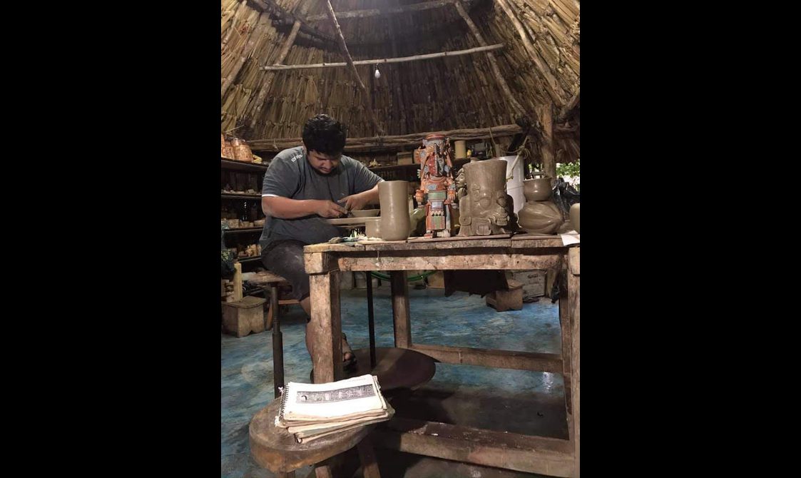 Man crafting pottery in a rustic workshop with a thatched roof, surrounded by clay artifacts and sculptures.