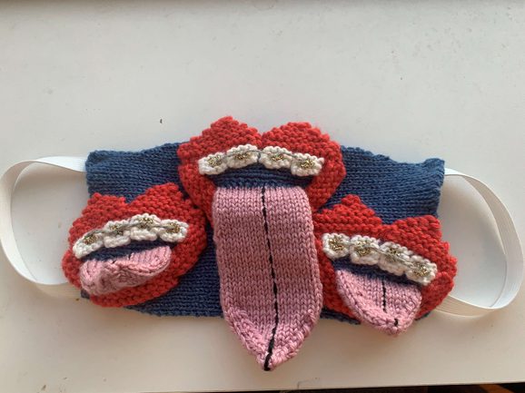 A knitted face mask featuring three mouths with braces on teeth and tounges sticking out.