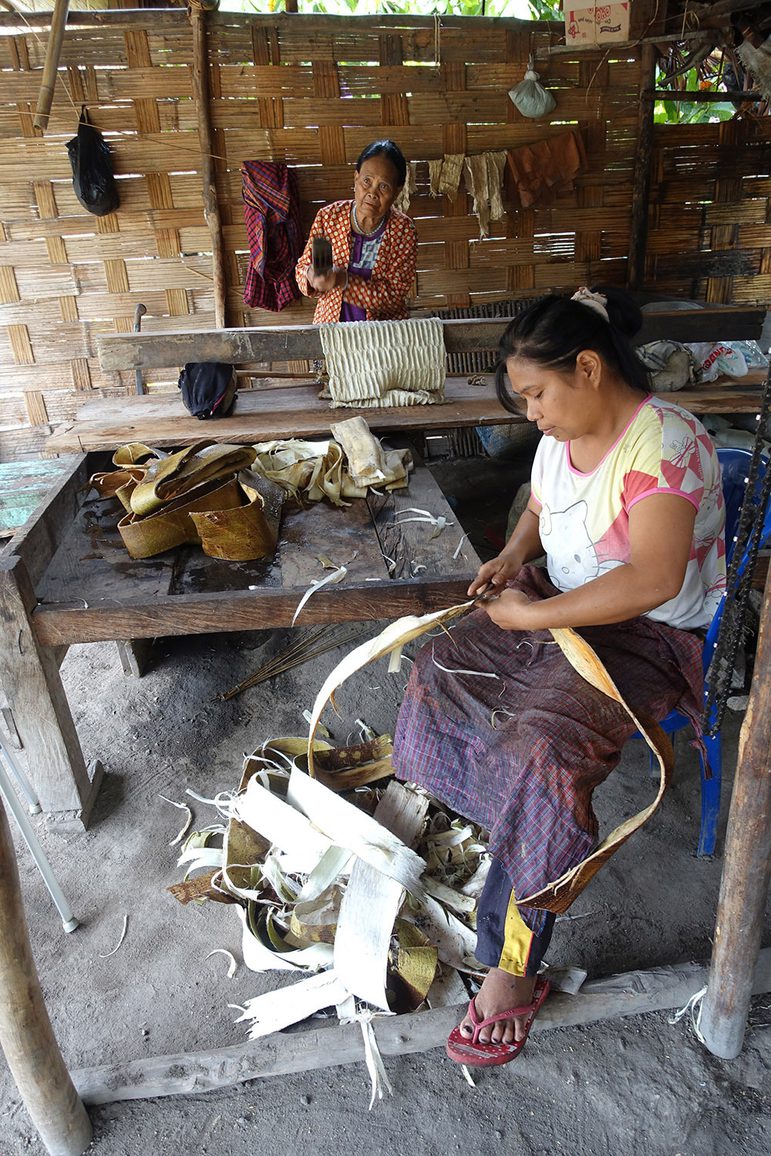 Two women making textiles from tree bark. The floor is dusty and there is a latticed woodwork wall behind them.